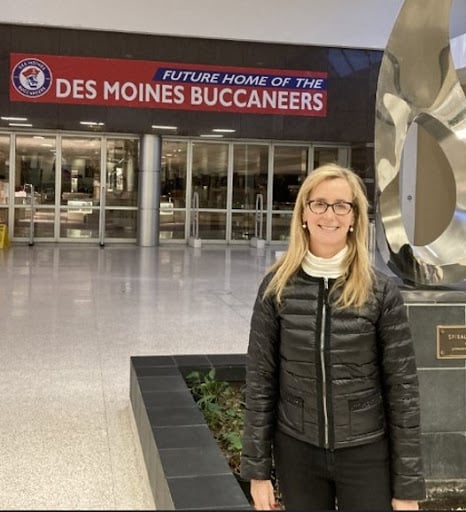 A photo of Elizabeth Holland in front of a sign that says "Future home of the Des Moines Buccaneers." She has long blond hair and wears a black coat and pants and black rimmed glasses