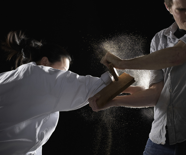 Young man holding plank, woman breaking plank with fist, side view